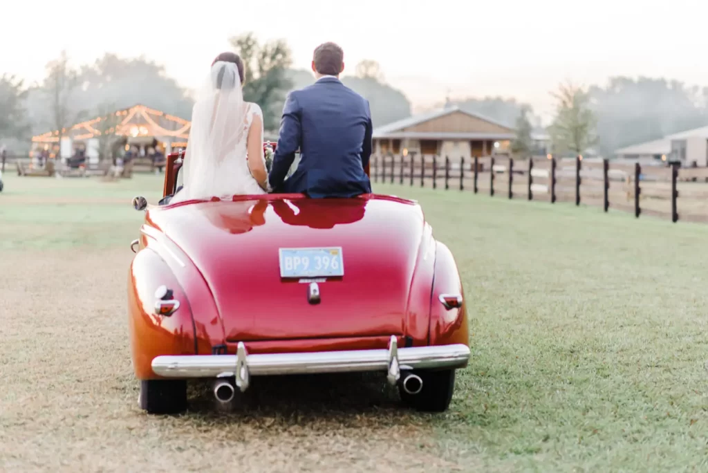 Bride and groom seated in the back of a red vintage car, leaving their wedding celebration.