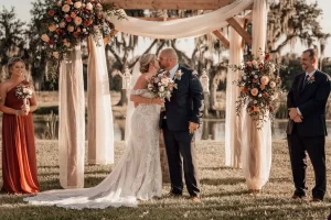 Bride and groom share a kiss under a floral archway during an outdoor wedding ceremony