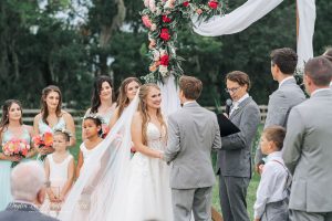 Bride and groom exchange vows under a floral arch during their outdoor wedding ceremony, surrounded by their wedding party and guests.